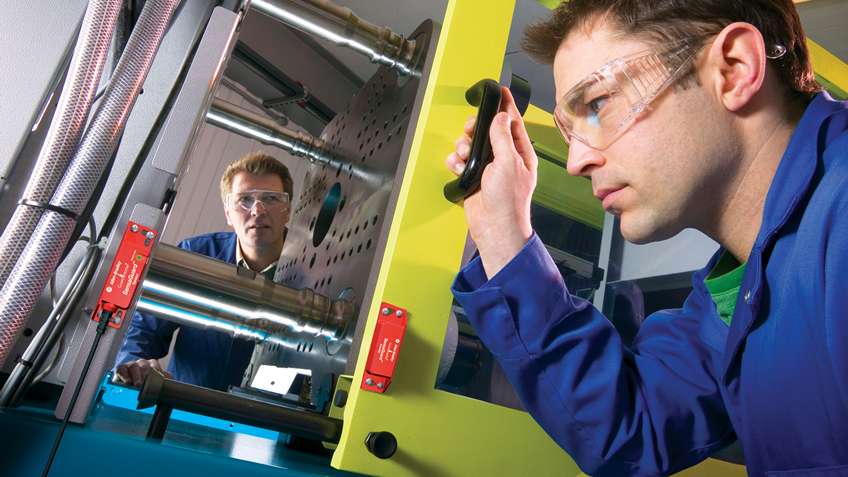 Two technicians in safety glasses inspecting a machine guard door fitted with safety interlock switches