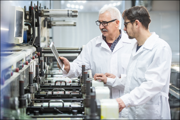 Two technicians in lab coats reviewing production data on a tablet at a roller-fed processing line
