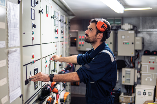 Technician operating switchgear controls at a motor control panel lineup while wearing hearing protection