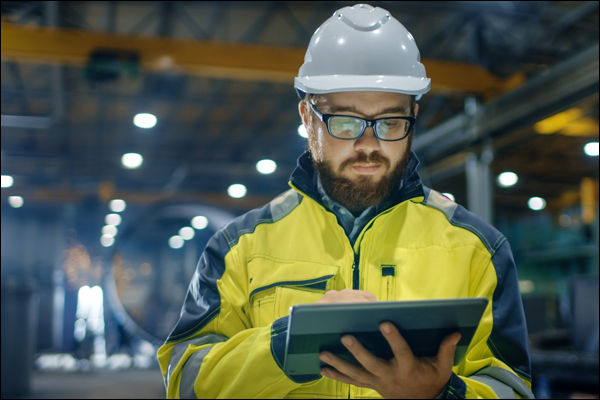 Worker in a hard hat, safety glasses, and high-visibility jacket using a tablet in an industrial facility
