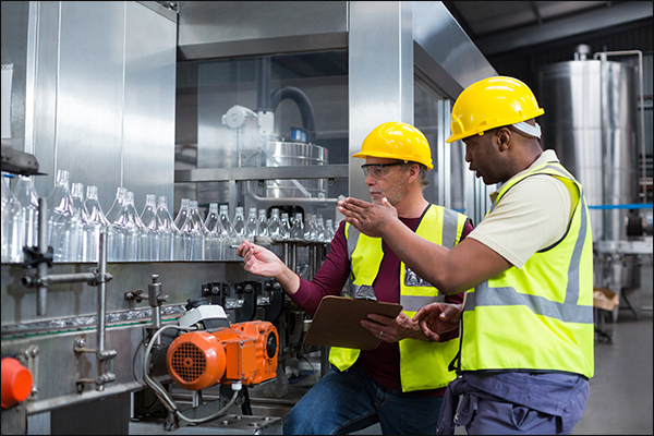 Two workers in hard hats and high-visibility vests inspecting bottles on a filling line with a tablet and clipboard