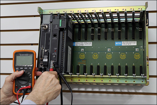 Technician testing an Allen-Bradley PLC chassis with a digital multimeter, processor and communication modules installed